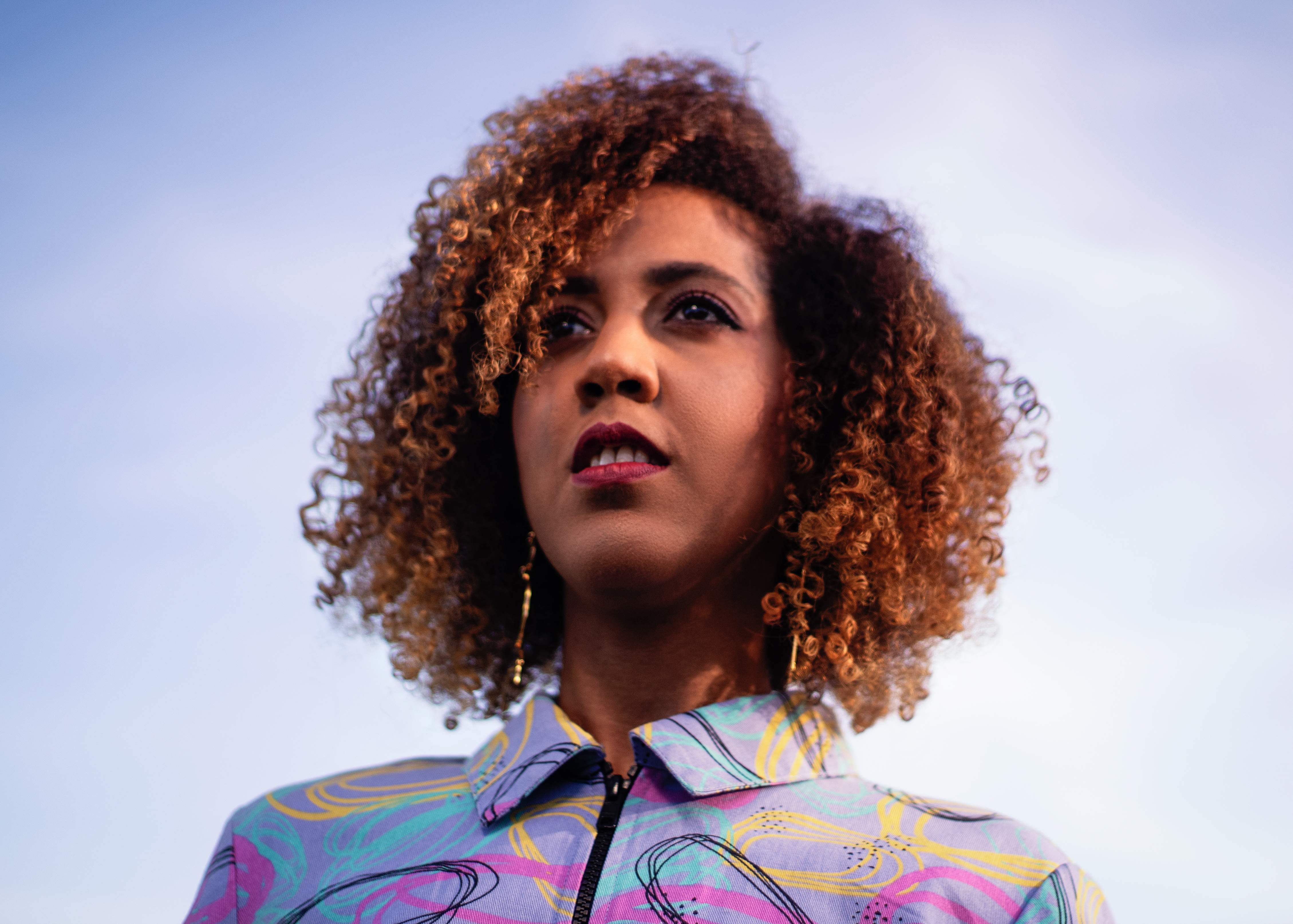 Woman with auburn afro curls, and magenta lipstick, wearing a brightly coloured geometric shirt and looking into the distance.