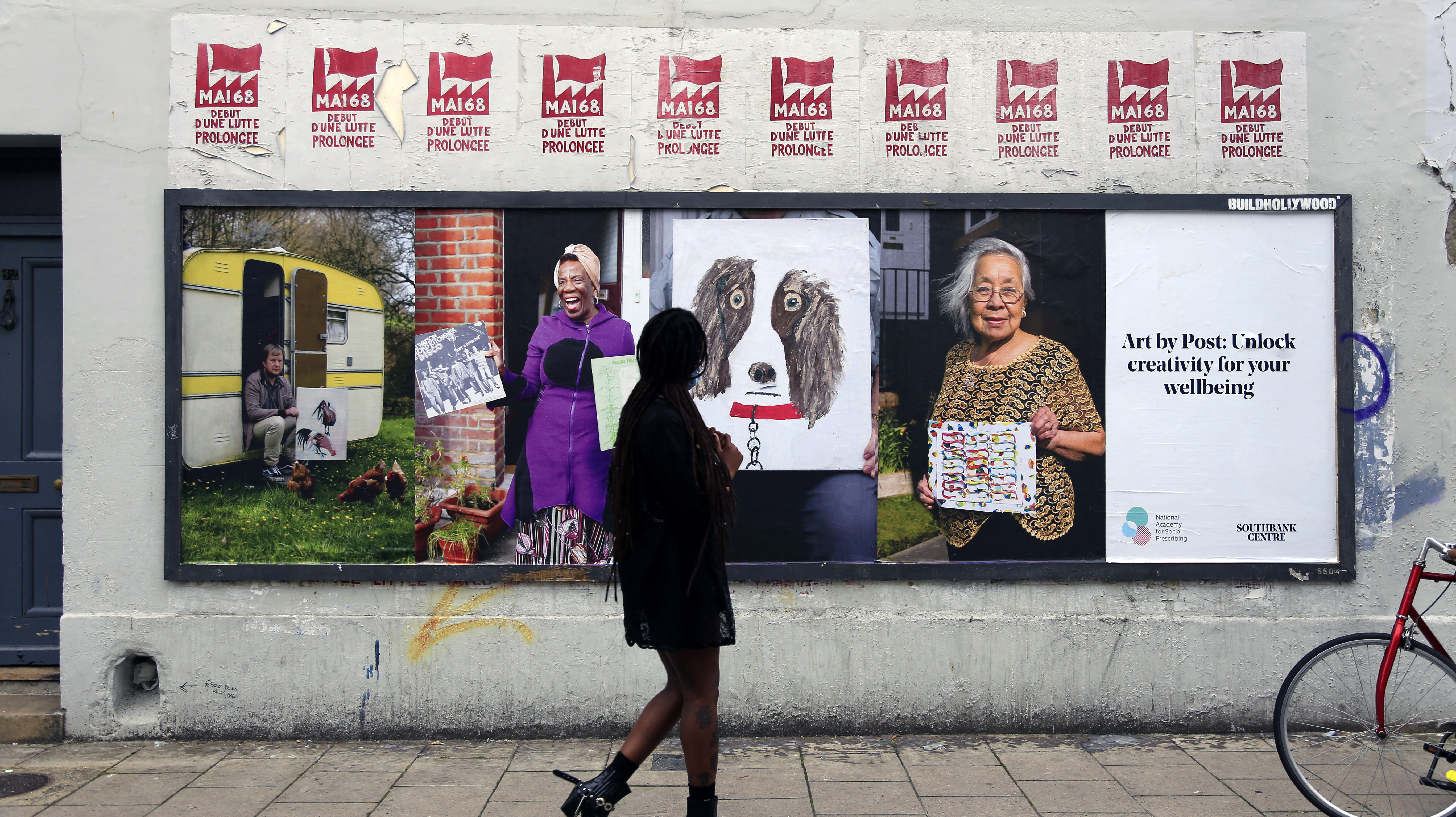 A woman walking past a billboard for Art by Post: Of Home and Hope featuring three artists holding their artwork