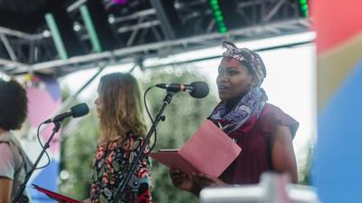 A woman holding a folder speaks into a microphone on stage alongside other performers.