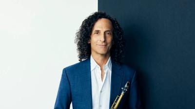 Kenny G in a blue suit holds a soprano saxophone, standing against a split white and dark blue background.