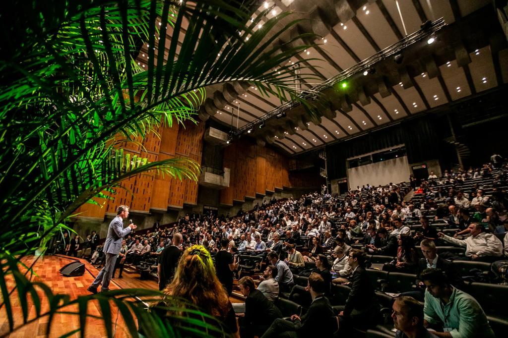 Person on stage holding a microphone in front of a full Queen Elizabeth auditorium. Plants in the foreground. 