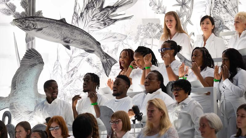 A diverse choir in white clothing sings passionately in front of a giant black and white sketch of a fish and flora.