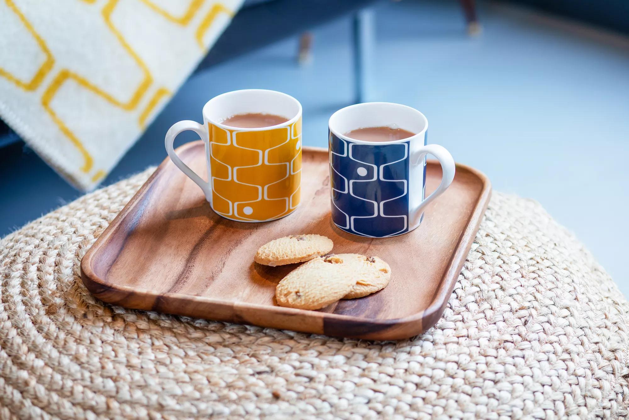 A wooden tray with cups of tea and chocolate chip cookies, balanced on a straw side table