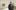 Artist Yoshitomo Nara – an older Japanese man with grey hair over which he wears a black baseball cap, and wearing a black hooded top, stands in his whitewalled artists studio which has paintings on the wall and various materials propped against the walls and at the room's edges.