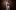 A person in a white suit and loose black shirt stood against a metal wall