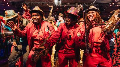 Musicians in matching red outfits and patterned hats smile while playing saxophones and flutes on a crowded dance floor.