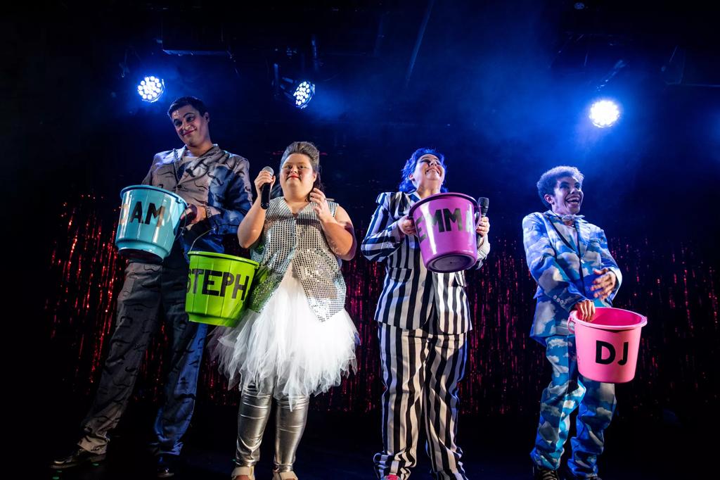 4 performers stand on a brightly lit stage with shimmering red angel hair streamers behind them. On the far left the man wears a grey suit with moustaches on and holds a blue bucket . Next to him a performer in a silver sequined waistcoat and tuille white skirt with silver leggings underneath holds a microphone and a green bucket. The third performer in line wears a white and black vertical striped suit, has blue hair and holds a microphone and purple bucket. On the far right the performer wears a sky blue suit with clouds on and holds a blue bucket. 