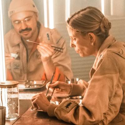 Two men sit on chairs playing music to a small group of people who stand and watch them.